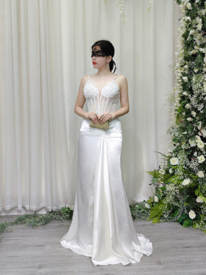 White beaded spaghetti strap mermaid gown with sheer corset bodice and satin skirt, worn with a lace mask and beaded clutch.