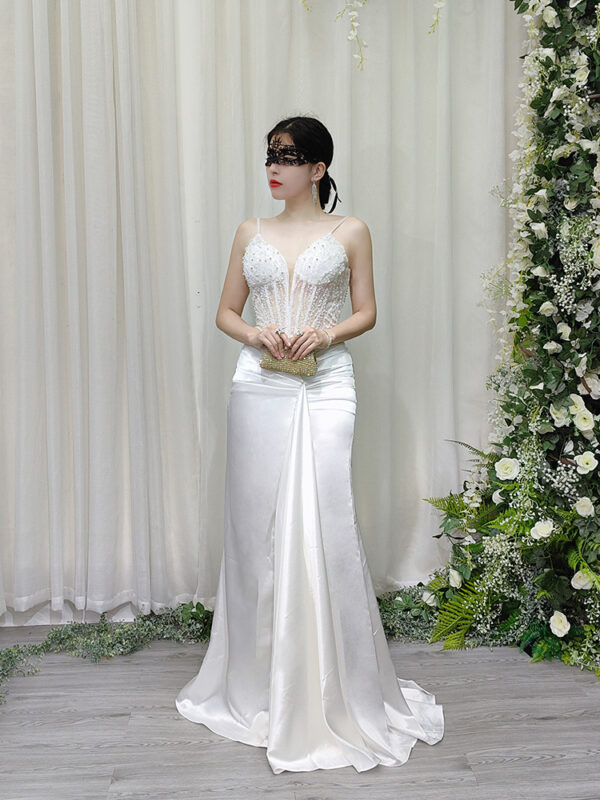 White beaded spaghetti strap mermaid gown with sheer corset bodice and satin skirt, worn with a lace mask and beaded clutch.