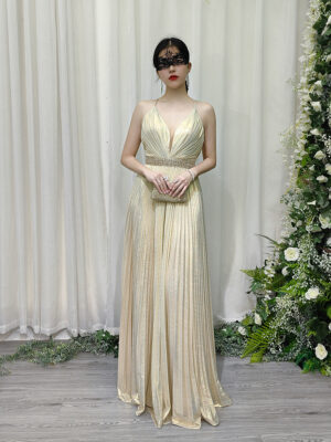 Champagne gold pleated halter gown with deep V-neck, beaded waistband, and flowing floor-length skirt, worn with a lace mask and beaded clutch.