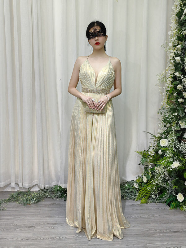Champagne gold pleated halter gown with deep V-neck, beaded waistband, and flowing floor-length skirt, worn with a lace mask and beaded clutch.
