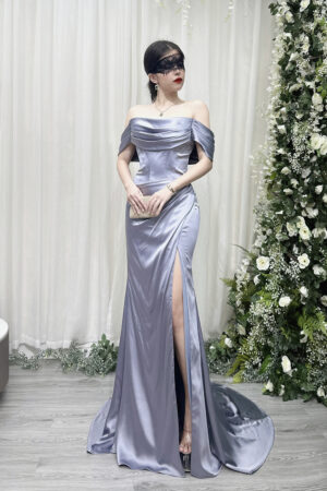 Dusty blue off-the-shoulder satin mermaid gown with corset bodice, high thigh slit, and sweeping train, worn with a lace mask and beaded clutch.