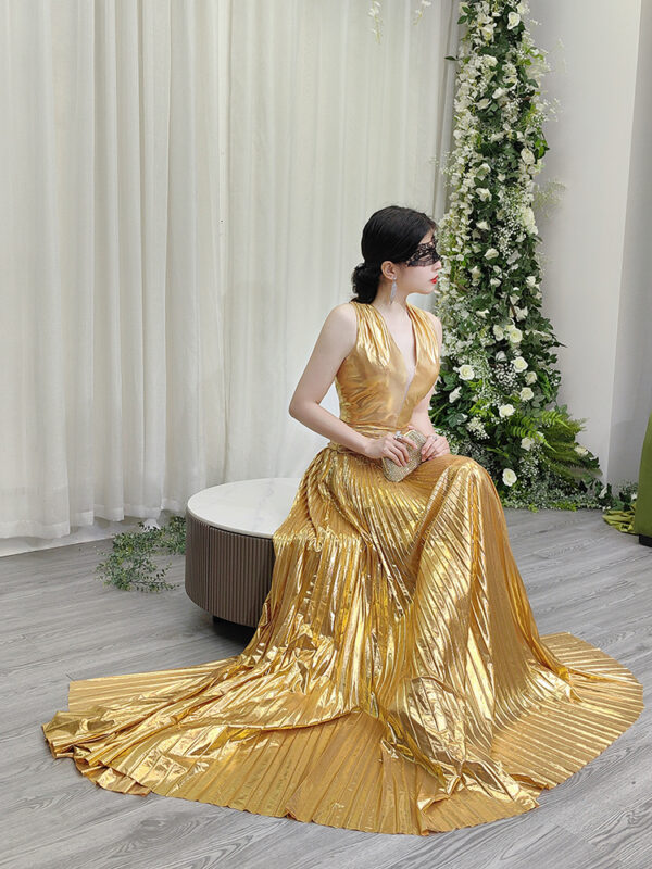 Modern gold metallic pleated gown with deep V-neck, ruched waist, high slit, and sweeping train, styled with a lace mask and beaded clutch.