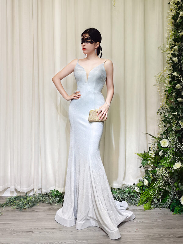 Elegant silver glitter mermaid evening gown with spaghetti straps and deep V-neck, worn by a woman in a black lace mask, holding a beaded clutch.